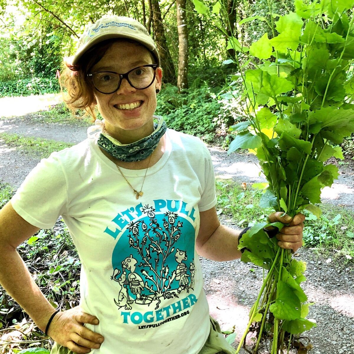 Michelle, a white woman with curly red hair and glasses, wears a baseball cap and holds a bunch of garlic mustard.