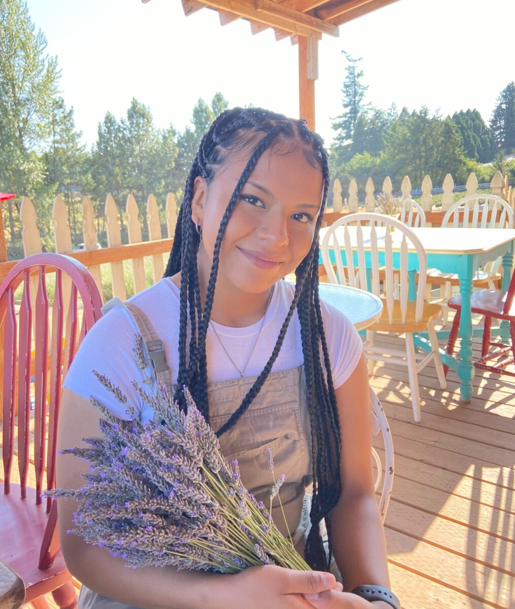 Picture of young woman with braids smiling and holding bouquet of lavender in a shady outdoor area.
