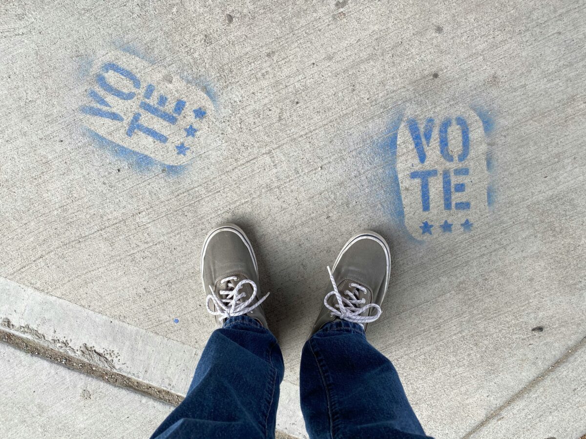 the word vote painted on the sidewalk with a person's shoes and legs standing over it