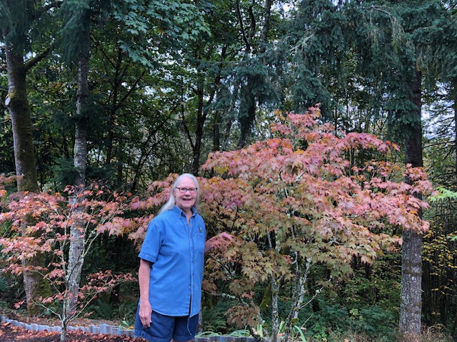 Dorinne, a white woman with long white hair, stands in front of a maple tree with glasses. She's smiling and wearing a blue shirt.