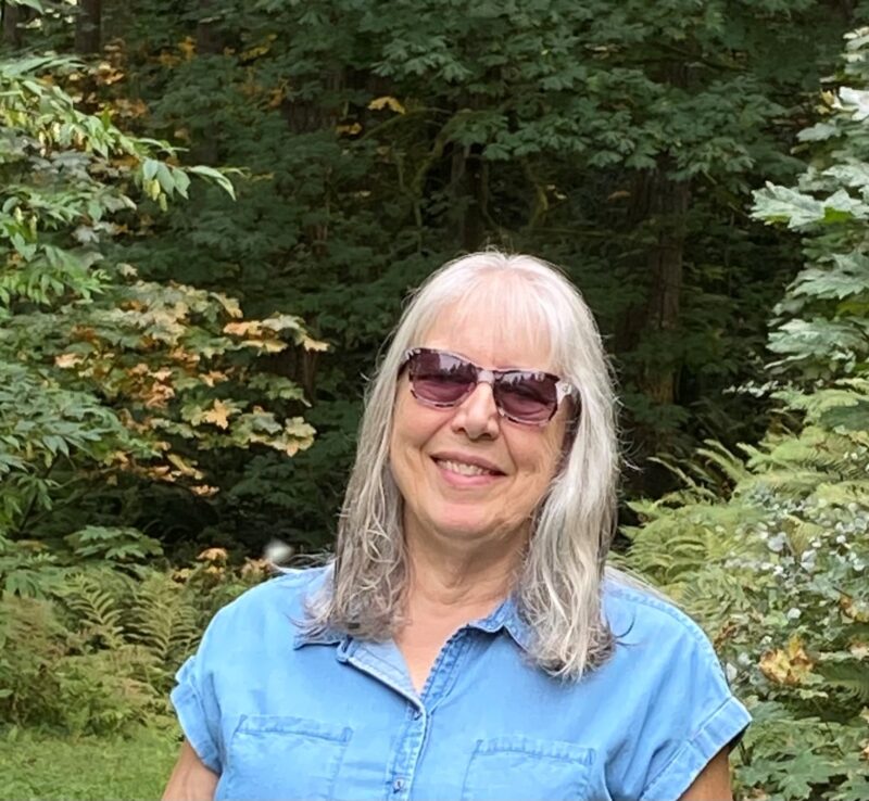 Rachael, a white woman with white hair and bangs, wears sunglasses and a blue shirt.