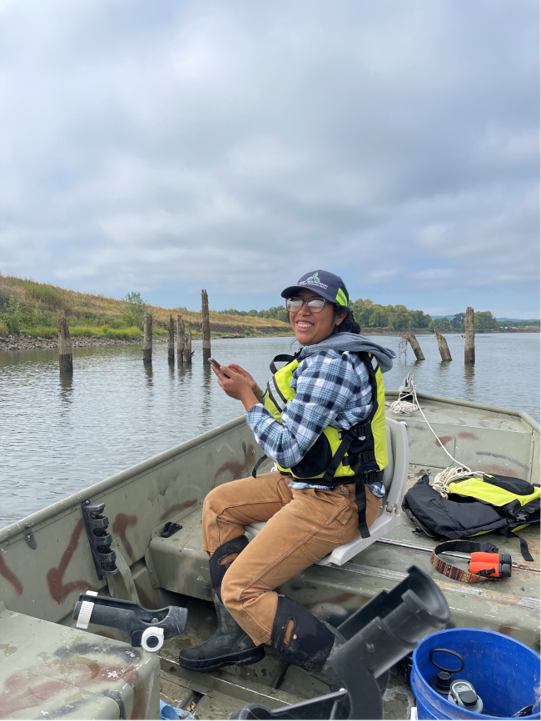 Martina sits in the boat holding her phone and smiling. She wears a plaid shirt and life vest.