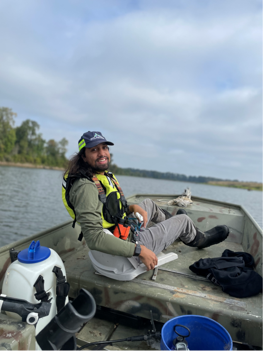 Shahbaz sits in the boat with water in the background. His dark hair is on his shoulder and he's smiling at the camera.