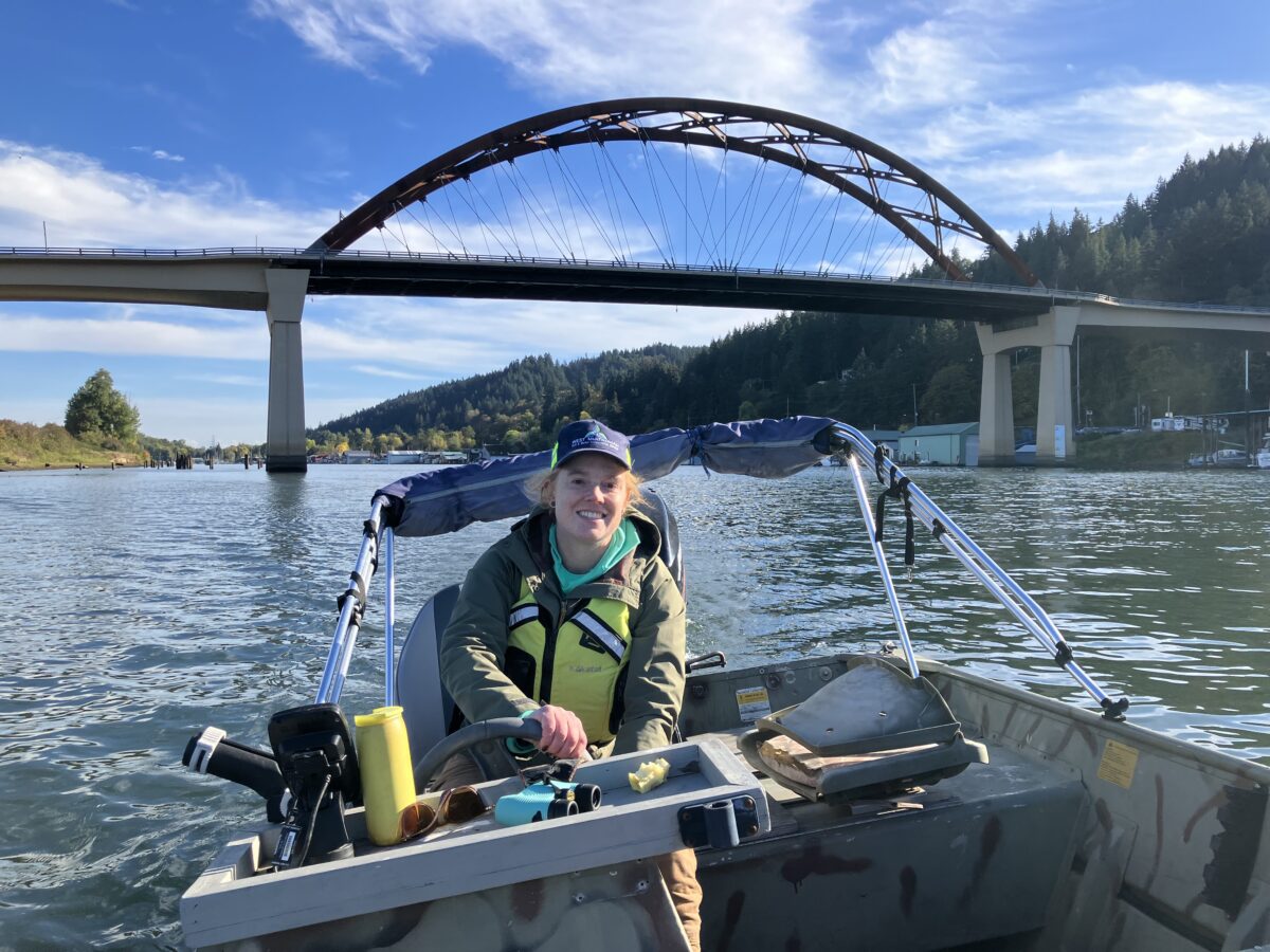 Michelle drives a boat under a bridge. She is a white woman smiling and wearing a yellow life jacket.