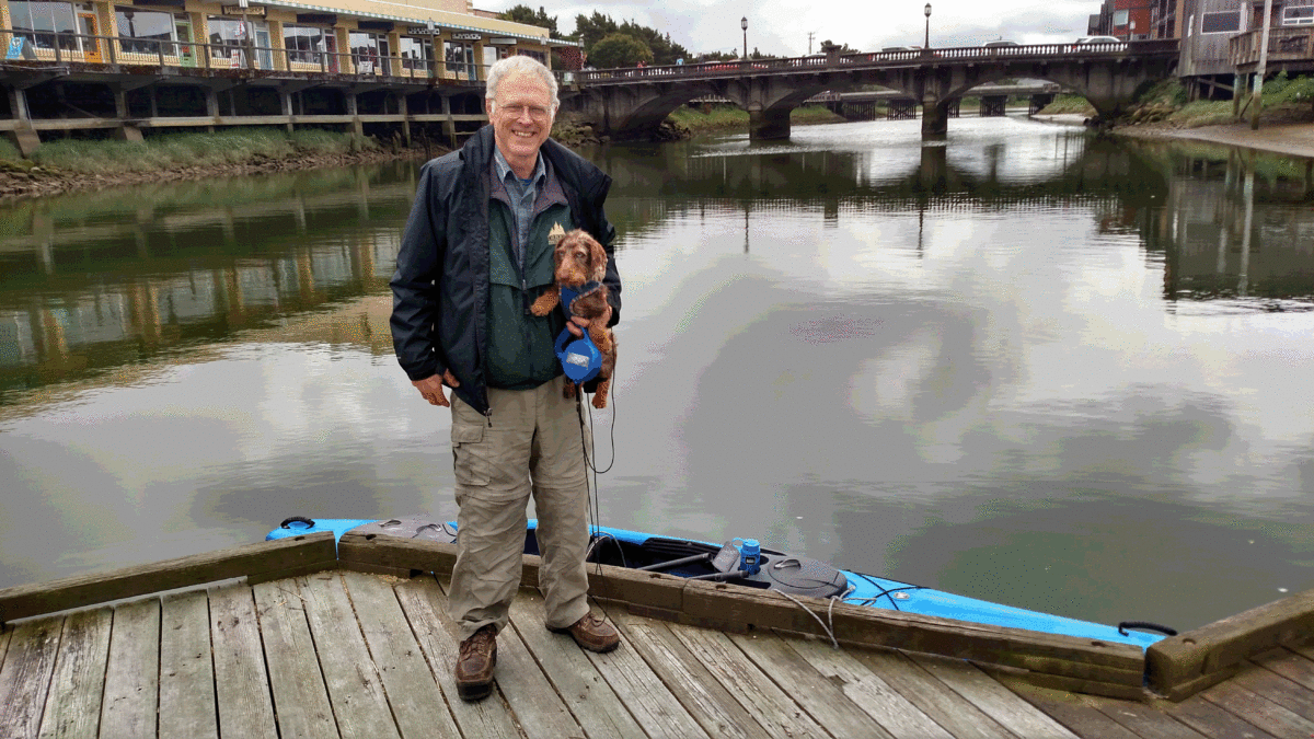 Older white man with white hair holds a small brown dog while smiling on a bridge in front of a large expanse of water.