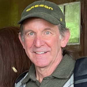 A close cropped picture of a white man with gray hair and a green hat smiling at the camera.