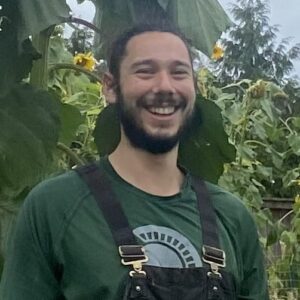 Josh, a white man with a dark beard and hair smiles in front of a sunflower garden.