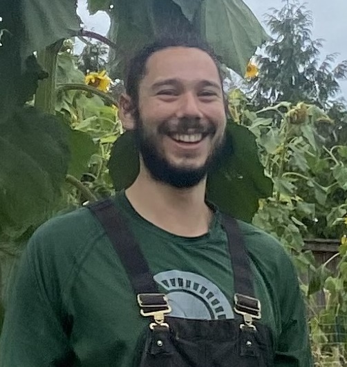 Josh, a white man with a dark beard and hair smiles in front of a sunflower garden.