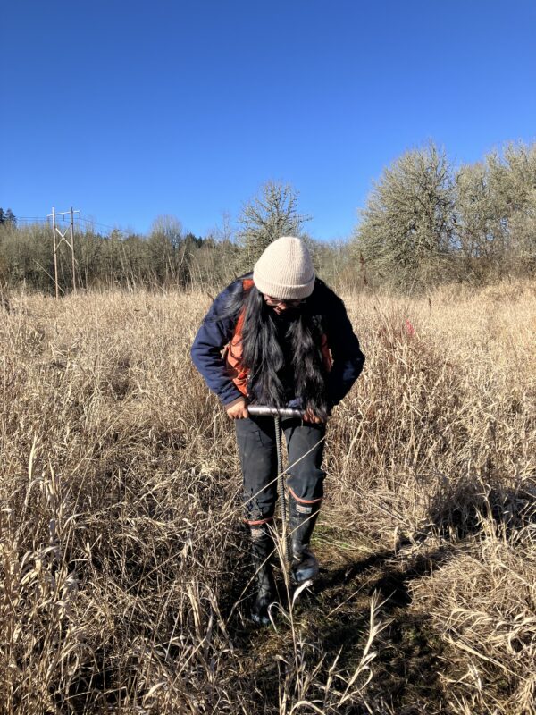 Martina looks down as she uses custom digging tool on property with tall grass