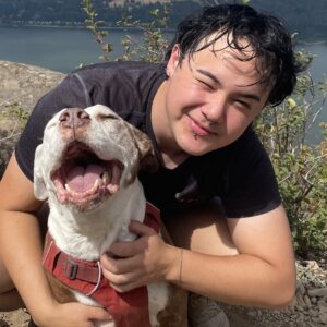 James has short dark hair and a black t shirt. He leans and squints at the camera while posing with a large white dog