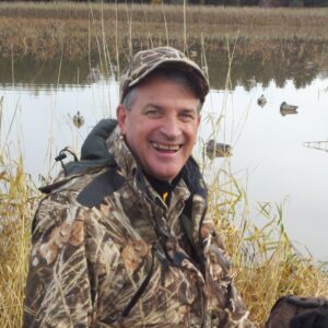 Roy kneels on the ground by a lake and a black dog, smiling and wearing a camouflage hat and jacket.