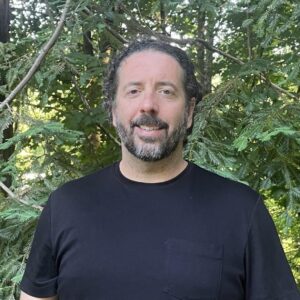 Jim Defuria, a white man with a black t-shirt and dark beard stands in the forest.