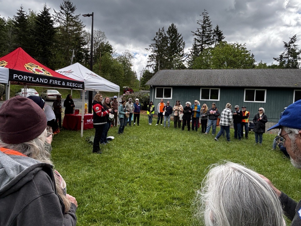A large crowd of people stands in a field in a circle, with tents and a building in the background.