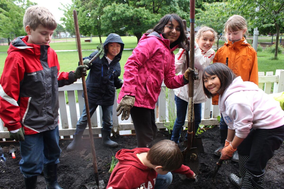 6 kids in raincoats with garden tools smile in a garden, with lots of green background