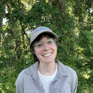 Gabrielle smiles at the camera in front of a leafy background wearing a beige hat and shirt.