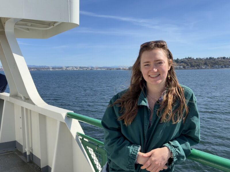Gillian smiles at the camera on the deck of a ferry with water and an island in the background.