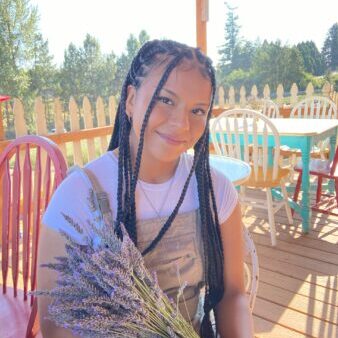 Picture of young woman with braids smiling and holding bouquet of lavender in a shady outdoor area.