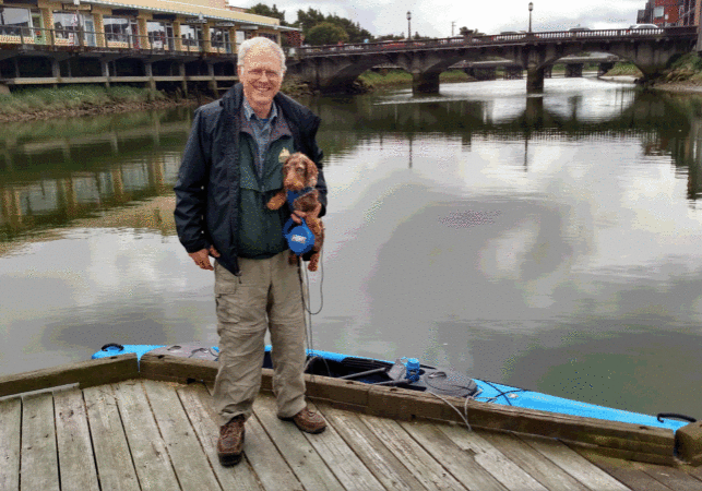 Older white man with white hair holds a small brown dog while smiling on a bridge in front of a large expanse of water.