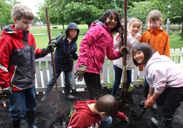 6 kids in raincoats with garden tools smile in a garden, with lots of green background