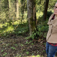 woman in brown sweater standing in forest with cleared ground in front of her