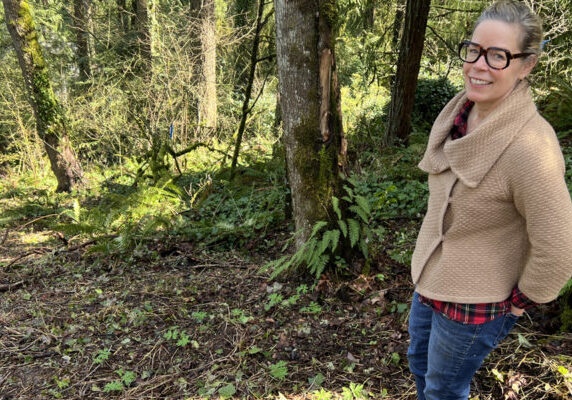 woman in brown sweater standing in forest with cleared ground in front of her