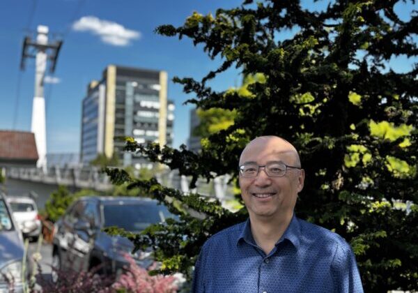 Sherman, a bald man in a blue shirt stands in front of a tree and the SW Portland skyline