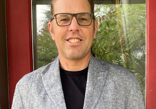 Jon stands smiling in a gray blazer and black glasses with a red door in the background.