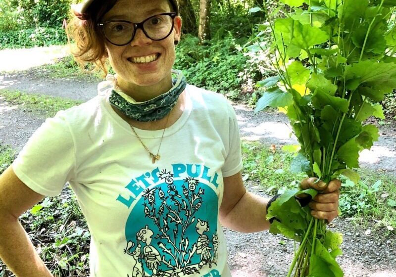 Michelle, a white woman with curly red hair and glasses, wears a baseball cap and holds a bunch of garlic mustard.