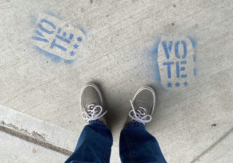 the word vote painted on the sidewalk with a person's shoes and legs standing over it