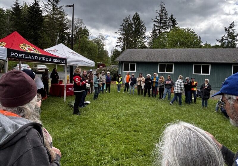 A large crowd of people stands in a field in a circle, with tents and a building in the background.